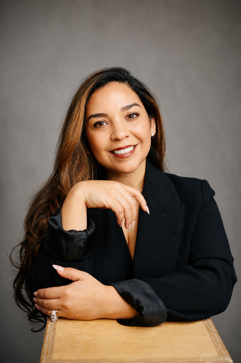 Portrait of a woman with long brown hair, smiling, wearing a black blazer, resting her chin on her hand with a ring visible. She is sitting at a table against a gray background.
