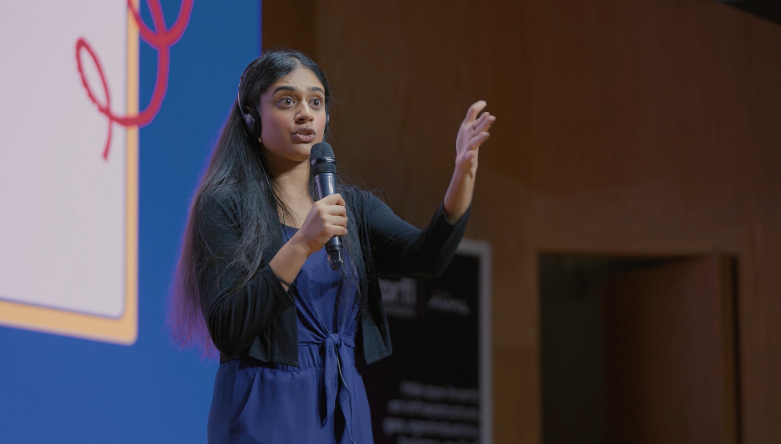 A woman speaking into a microphone during a presentation, wearing a blue dress and a black blazer, with a large colorful display behind her.
