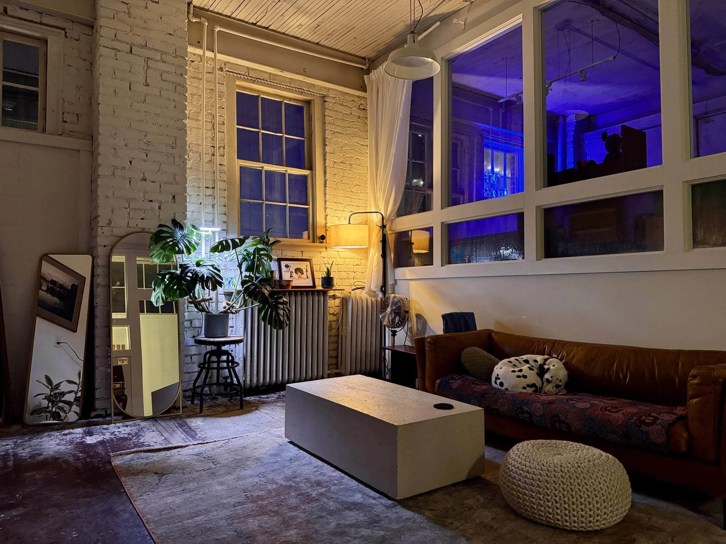 A cozy living room with exposed white brick walls and large windows. There is a brown leather couch with a Dalmatian-patterned cushion, a round knitted ottoman, and a rectangular concrete coffee table. A floor lamp, a potted monstera plant on a stand, and framed pictures on the radiator enhance the space. The room is softly lit, with some blue lighting visible outside the windows.