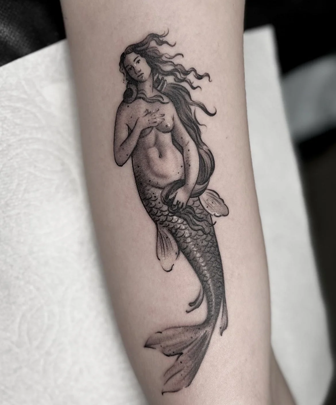 Tattoo of a mermaid with long flowing hair, holding her chest with one hand and the other resting on her tail, in black and gray ink.