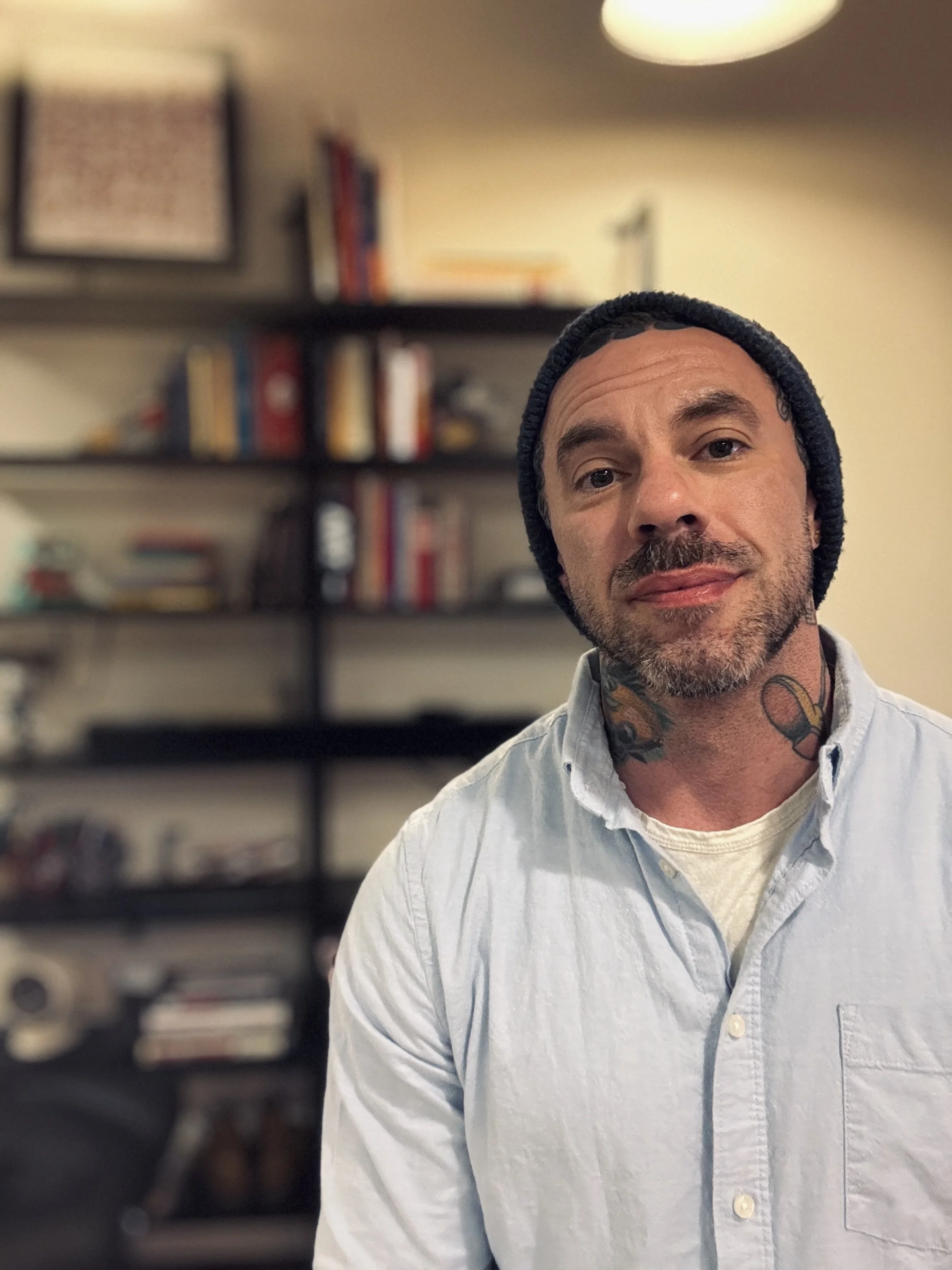 A man wearing a beanie and a light-colored shirt, standing in front of a bookshelf with various books and decorative items in a room with warm lighting.
