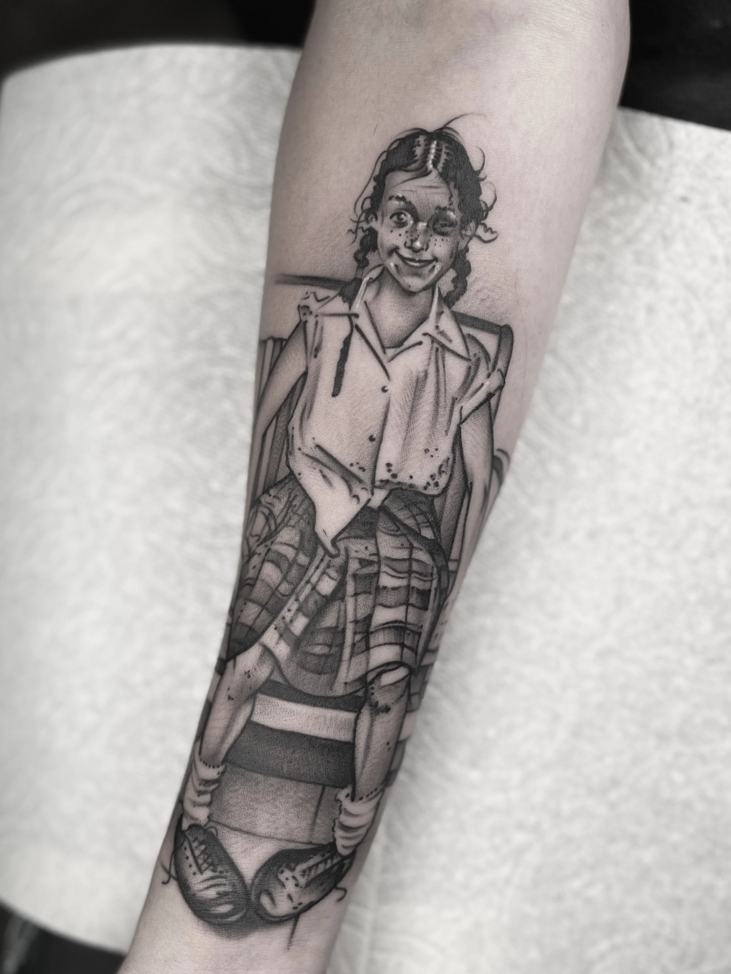 Black and gray tattoo of a girl with glasses winking and smiling, sitting on a school desk, with crossed legs, wearing a school uniform and sneakers.