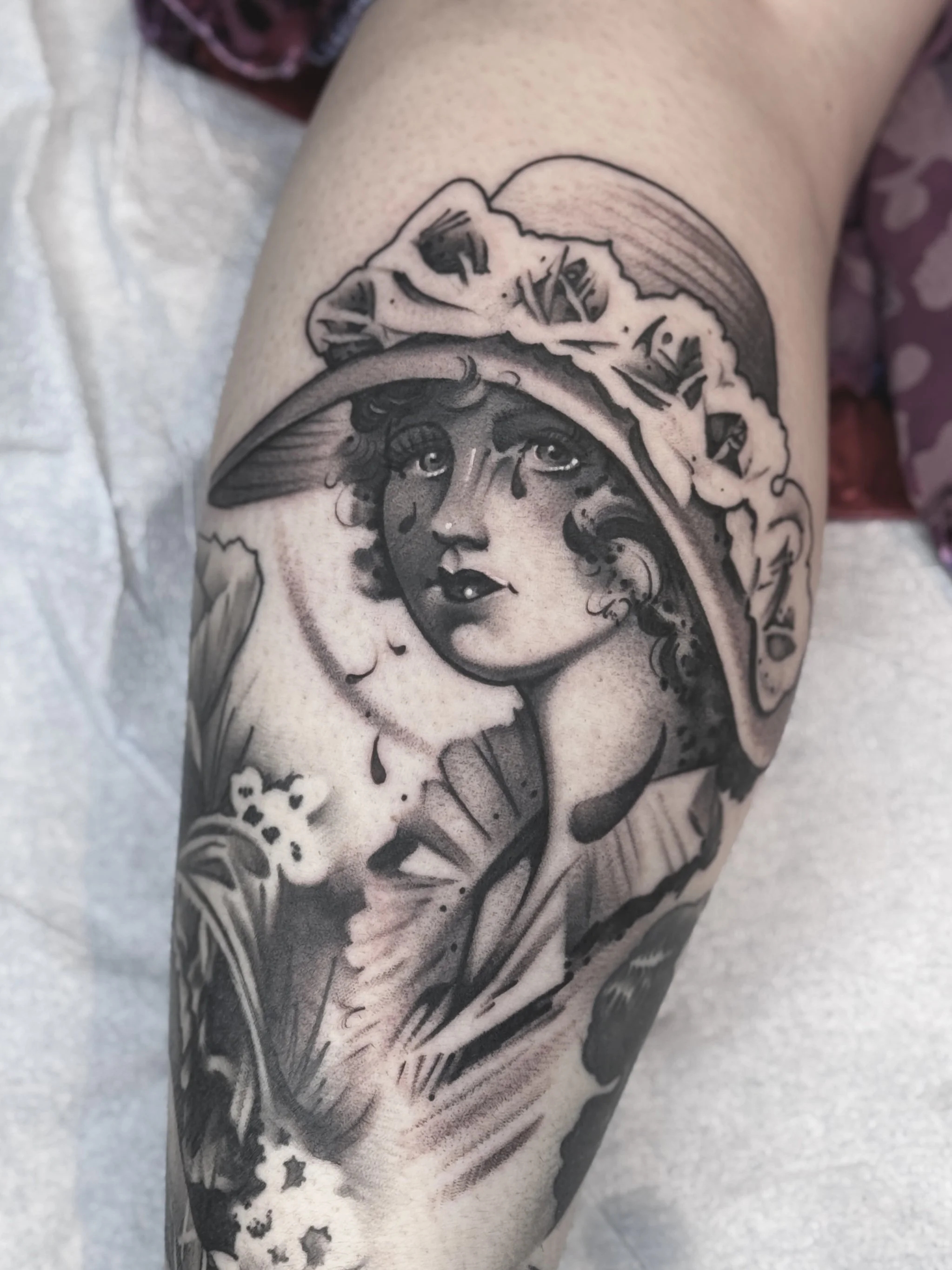 A black and gray tattoo of a woman's face wearing a large, wide-brimmed hat with a floral decoration. The woman has curly hair, expressive eyes with tears, and a subtle, serious expression.