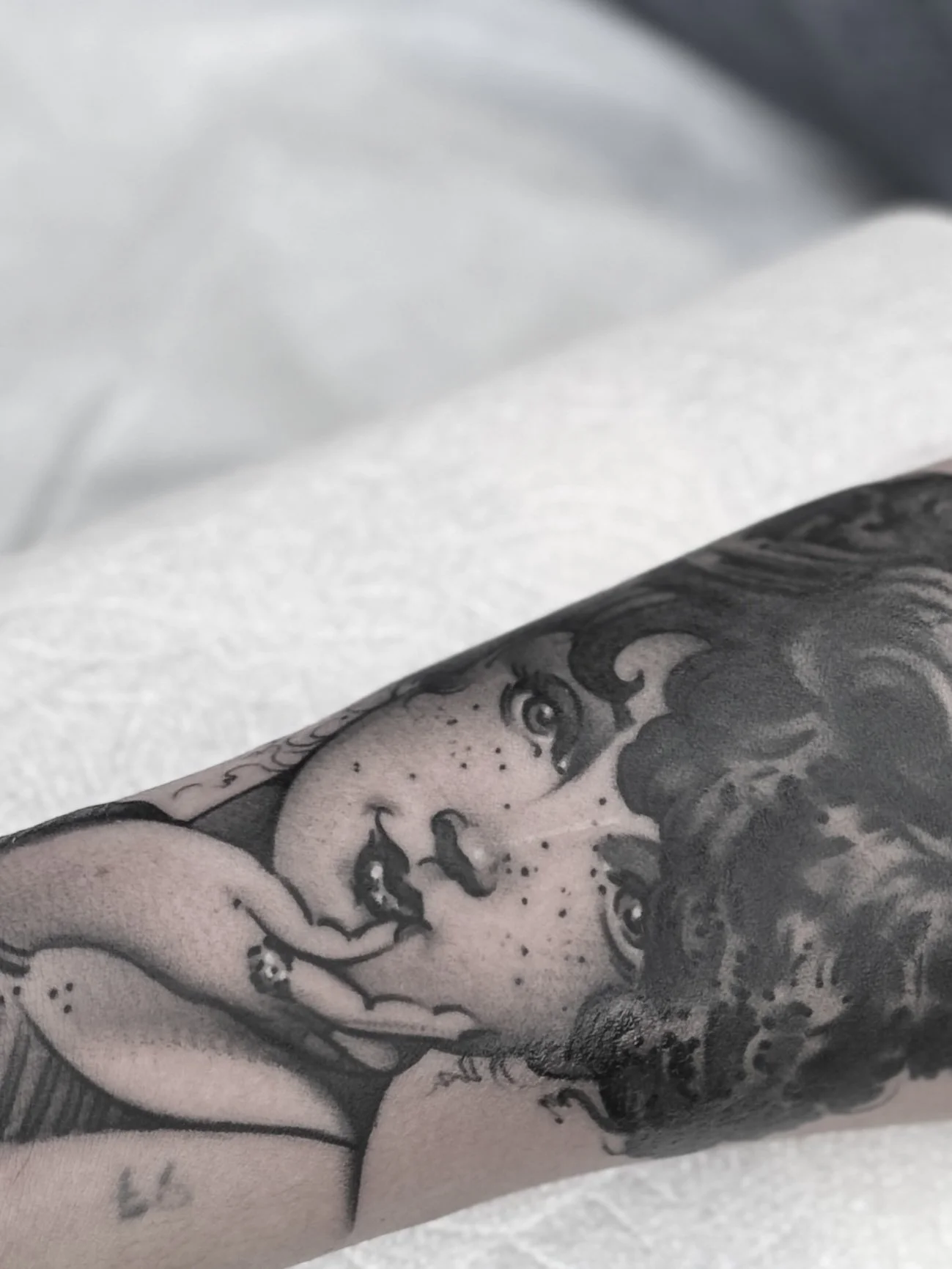 Close-up of a black and gray tattoo of a woman with curly hair, freckles, and makeup, with her finger near her lips.