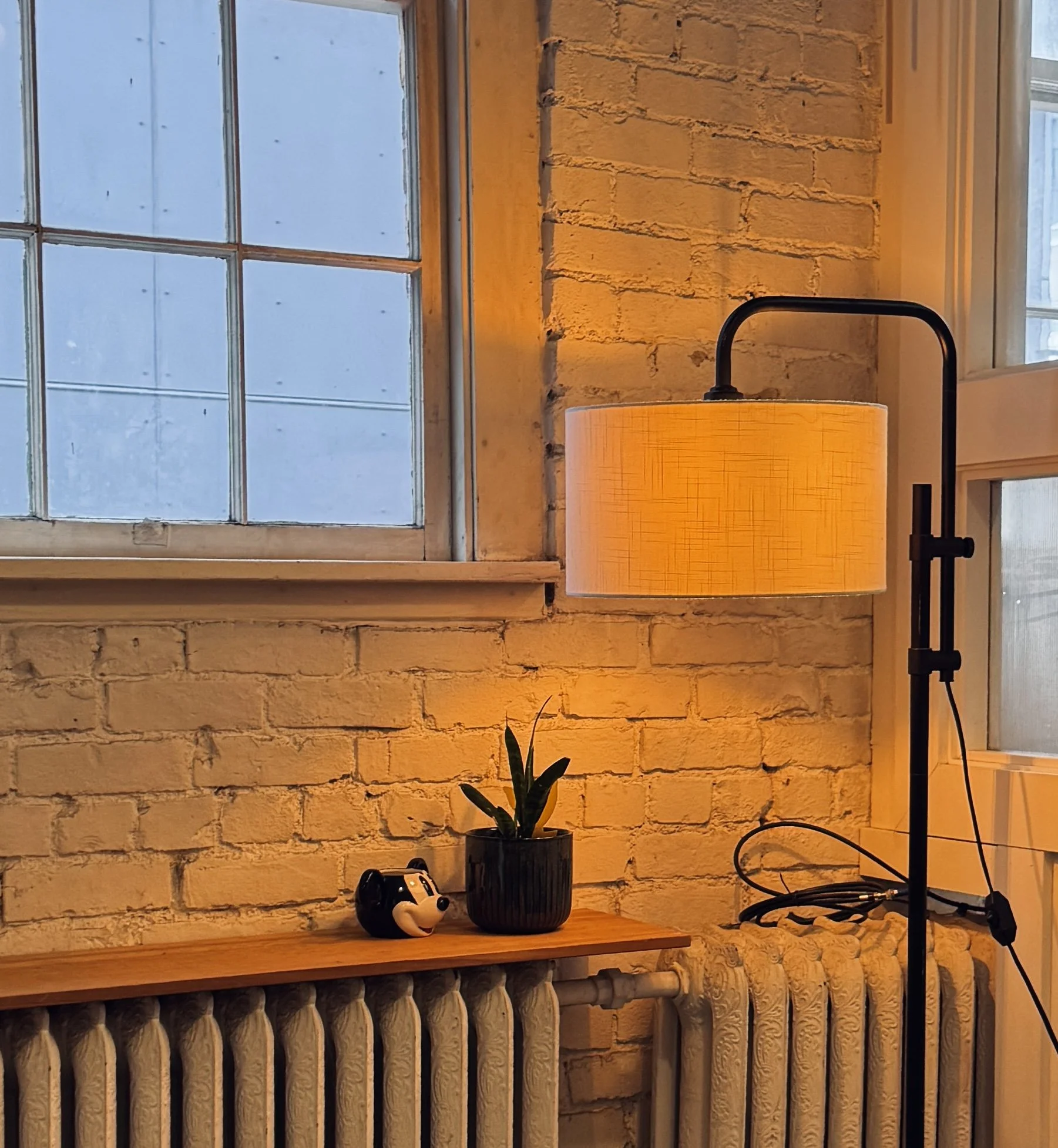 A cozy interior corner featuring a white brick wall, a wooden shelf with a small black pot holding a green succulent, a panda-shaped mug, and a tall floor lamp with a beige lampshade emitting warm light next to two windows.