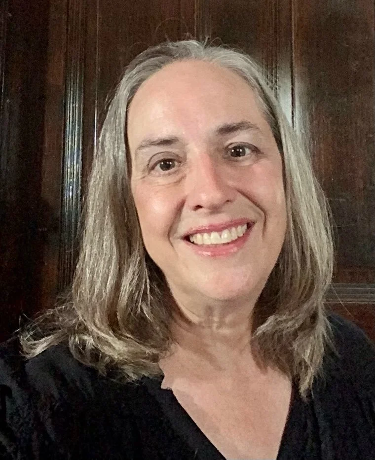 Portrait of a smiling middle-aged woman with long gray hair, wearing a blue sweater, against a plain light background.