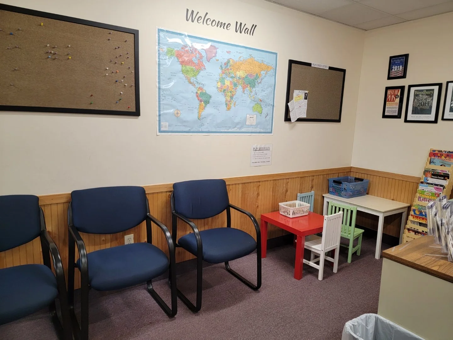 Waiting area with four navy blue chairs, small colorful tables, and a child's desk with three chairs. Wall decor includes a world map, framed certificates, and bulletin boards, with a magazine rack on the right.