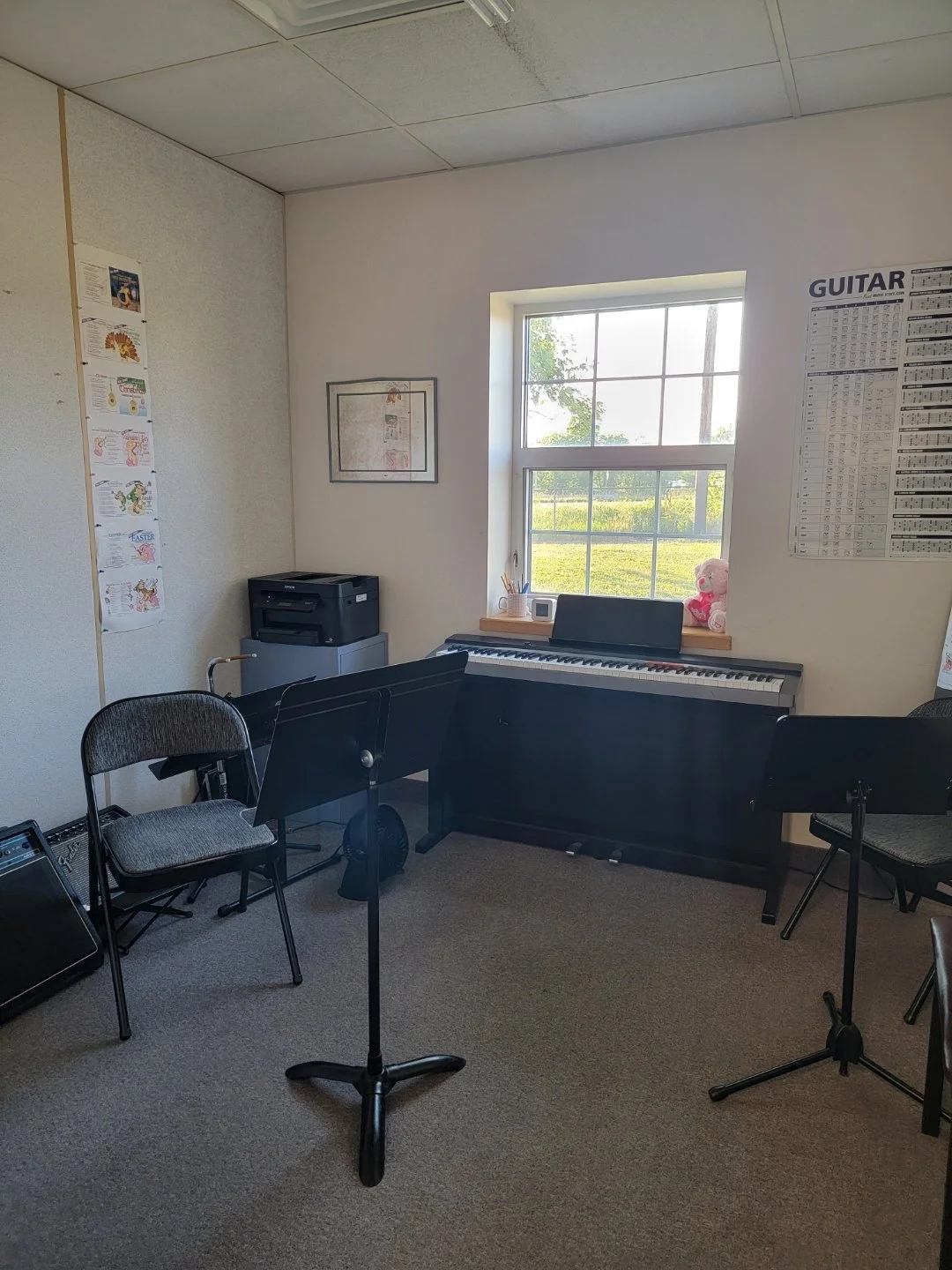 Music classroom with a piano placed beneath a window, chairs with music stands, a printer on a small table, framed picture on the wall, posters on the wall, and a large chart labeled "GUITAR" on the wall.