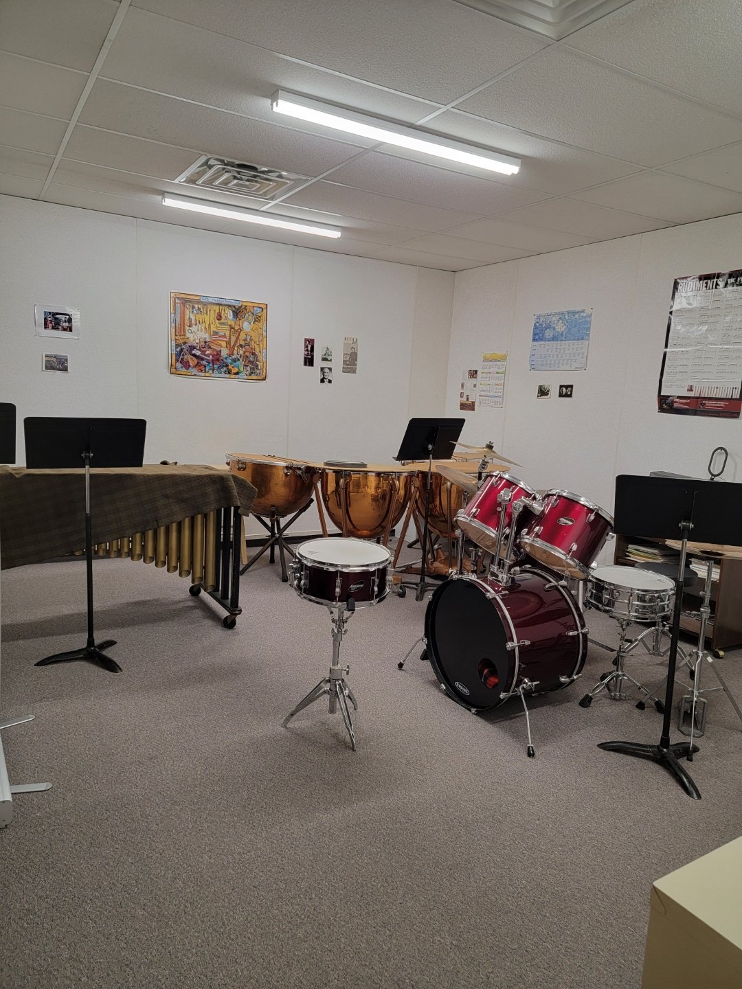 Music practice room with percussion instruments including conga drums, a drum set, and a marimba, along with music stands and posters on the white walls.