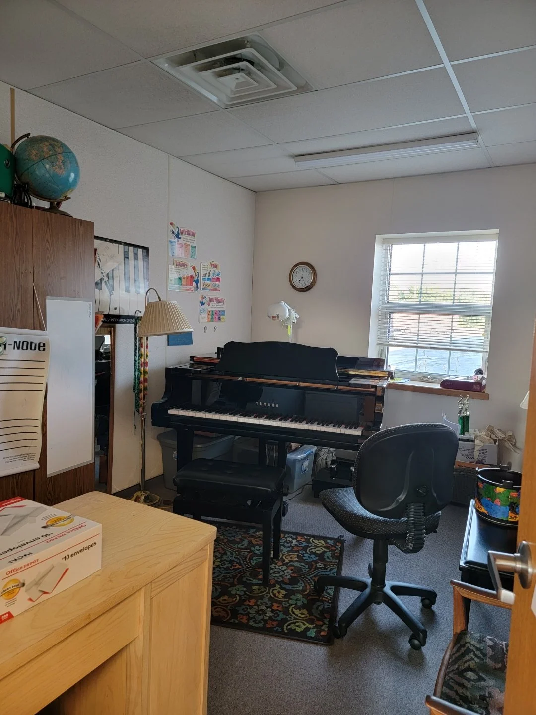 A small office or music room with a Yamaha upright piano, office chair, window, clock, globe, and various office supplies and decorations.