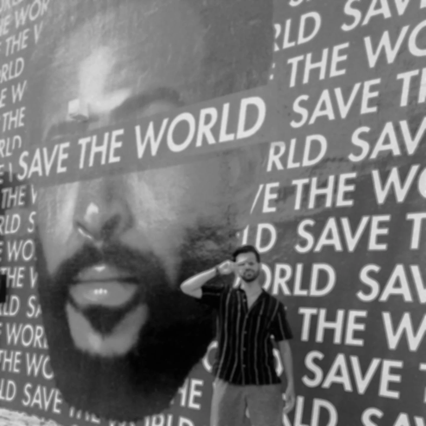 A man with glasses and a striped suit standing in front of a large wall art featuring a superimposed, close-up black and white face of a woman with the text 'Save the World'. The background pattern repeats the 'Save the World' text multiple times.