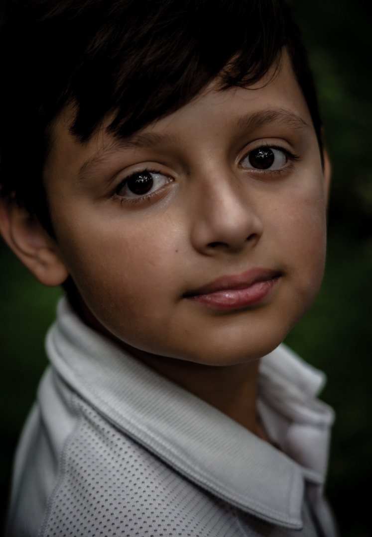 A young boy with short dark hair and light brown skin, looking directly at the camera with a gentle expression, wearing a white collared shirt, background blurred with greenery.