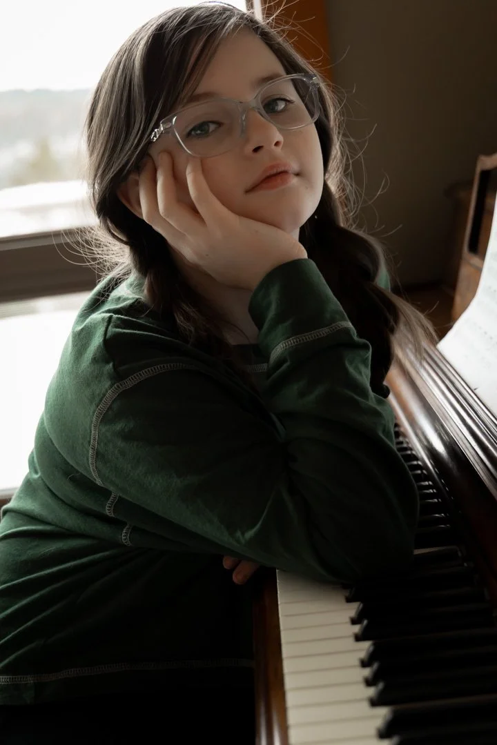 A young girl with glasses sitting at a piano, resting her head on her hand, looking at the camera.