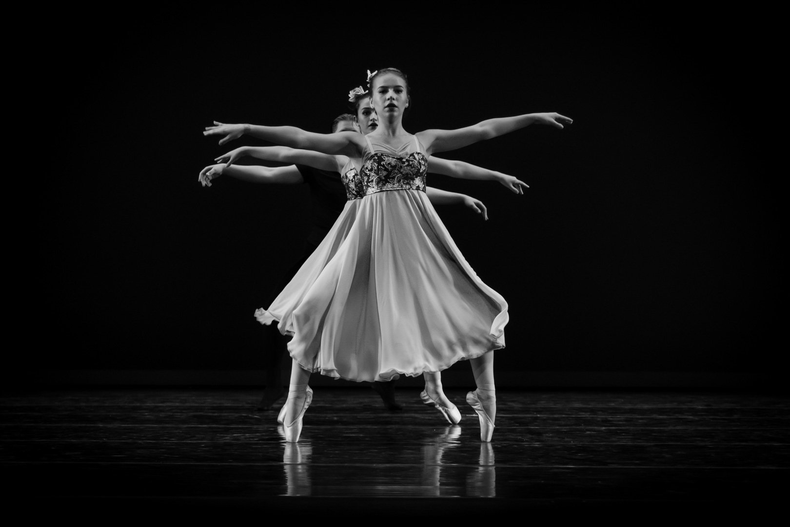 Black and white photo of three ballet dancers performing on stage, with one dancer in the front and two behind, all wearing elegant costumes and pointe shoes.
