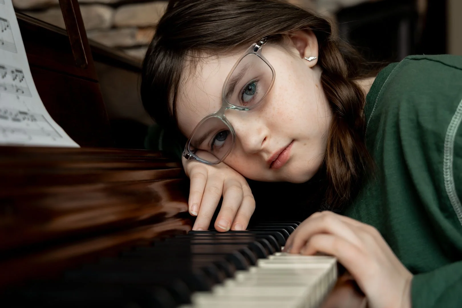 Young girl with glasses resting her head on a piano, playing keys with a contemplative expression.