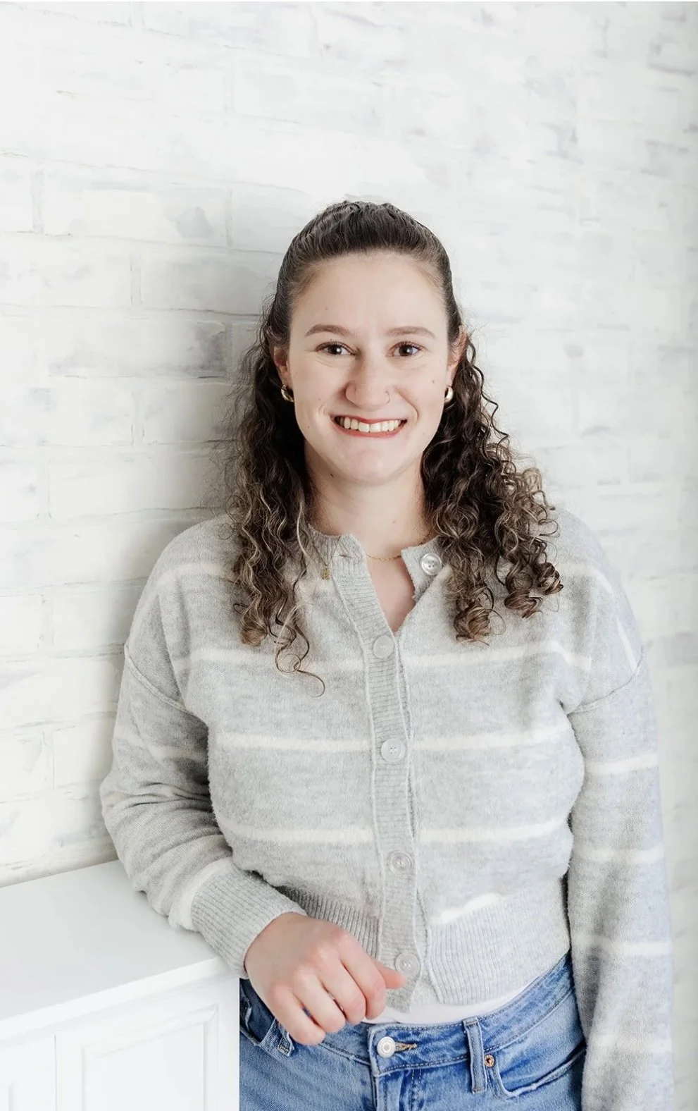A young woman with curly brown hair smiling, wearing a light gray striped sweater and blue jeans, standing against a white brick wall.