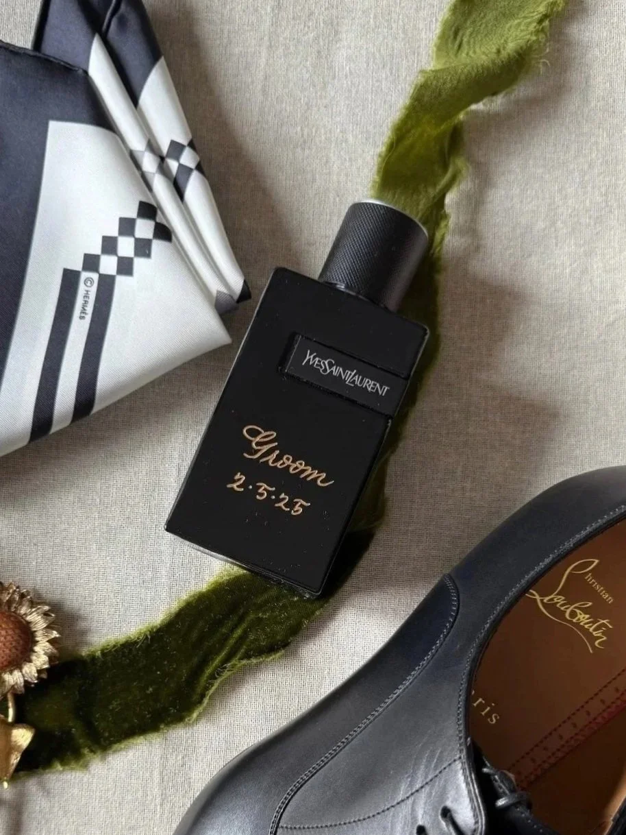 A black Yves Saint Laurent perfume bottle with personalized gold writing, a black Christian Louboutin shoe, and parts of a white and black patterned bag are visible on a light-colored surface.
