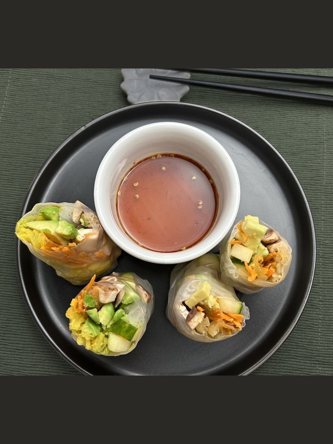Black plate with 4 Asian Spring Rolls with mushrooms, raw vegetables and fermented sauerkraut and a container of red dipping sauce - top view of the plate.