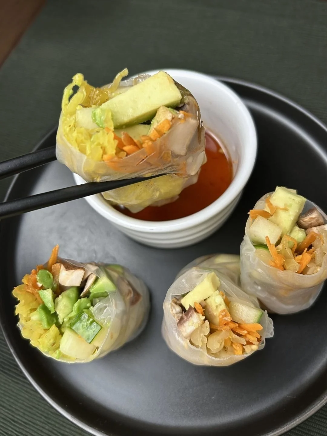 Black plate with 4 Asian Spring Rolls with mushrooms, raw vegetables and fermented sauerkraut and a container of red dipping sauce - side angle view of the plate with chopsticks holding one roll over sauce.