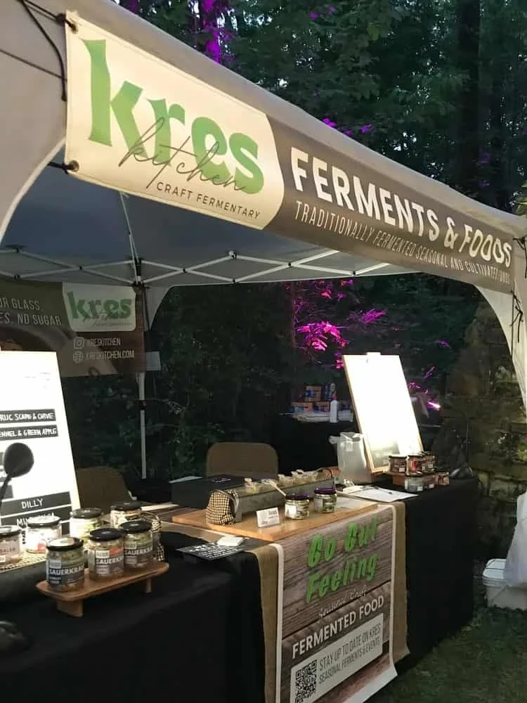 Kres Kitchen's professional sales booth set-up at an evening outdoor market with a lush green backdrop and florescent lights. The booth table is covered with a black table cloth and holds Kres Kitchen display jars for multiple sauerkraut varieties.