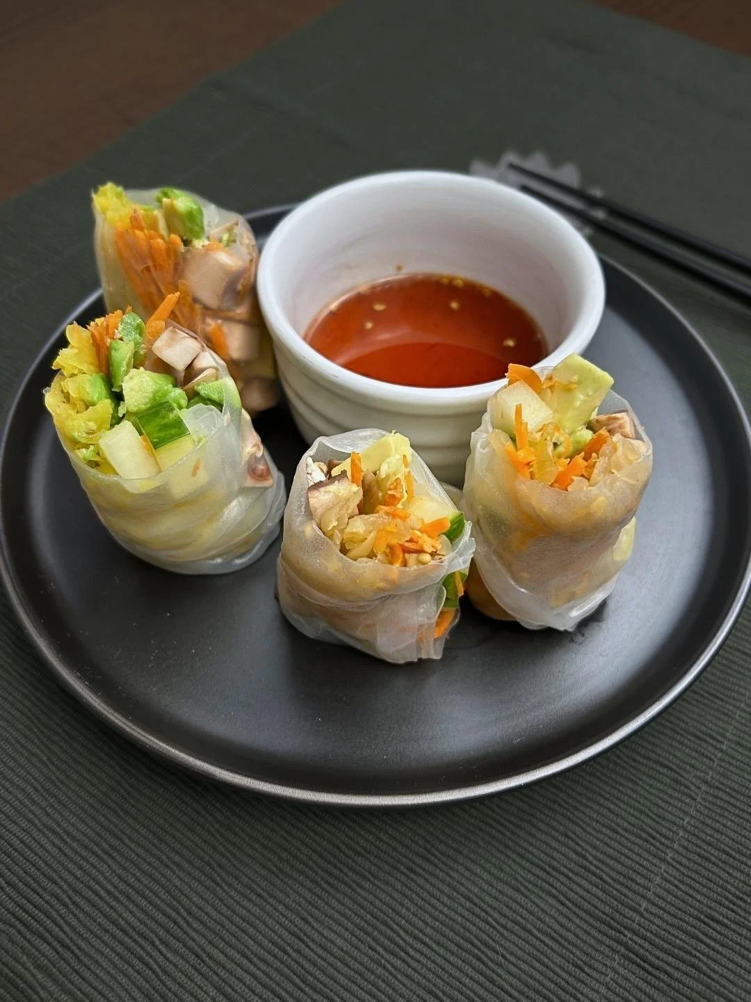 Black plate with 4 Asian Spring Rolls with mushrooms, raw vegetables and fermented sauerkraut and a container of red dipping sauce - side angle view of the plate.