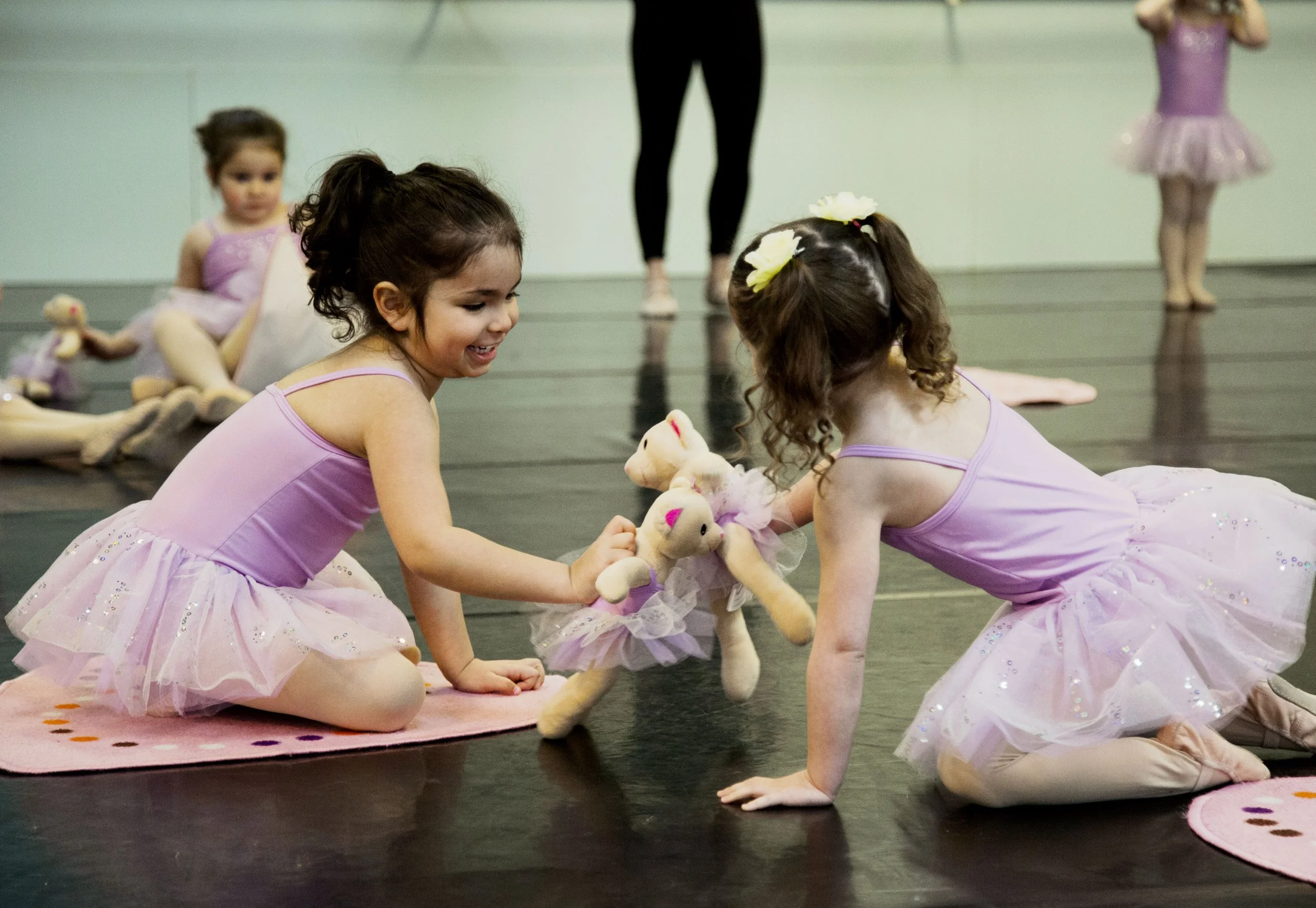 Young ballet dancers in pink tutus sitting on the floor during a practice, holding teddy bears, with a ballet studio's mirror and instructor in the background.