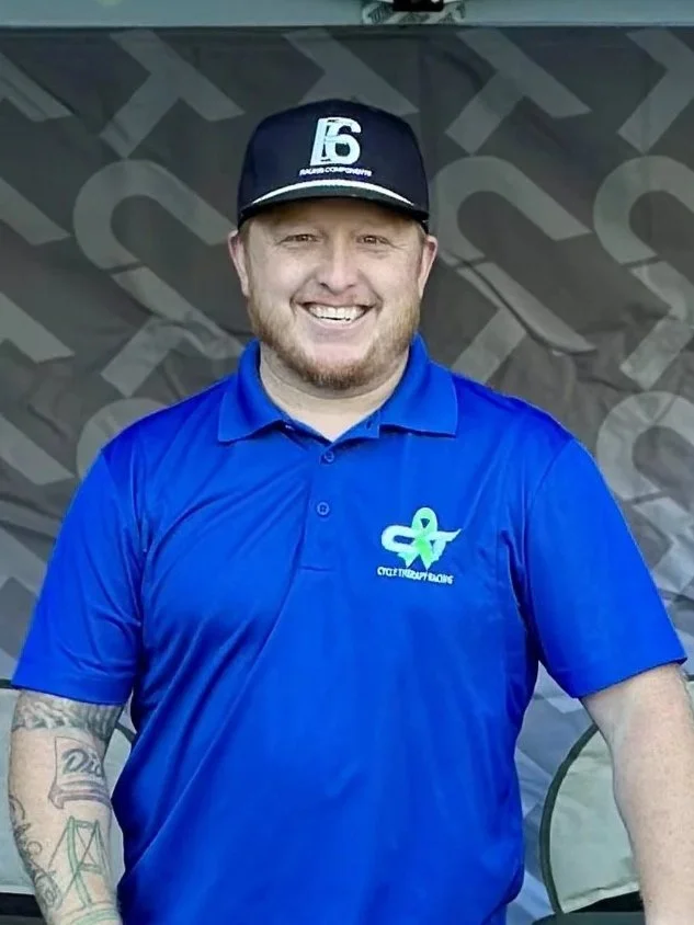 A man with light skin, short light brown hair, and a light beard, smiling and wearing a blue polo shirt with a logo on it, standing against a plain beige wall.