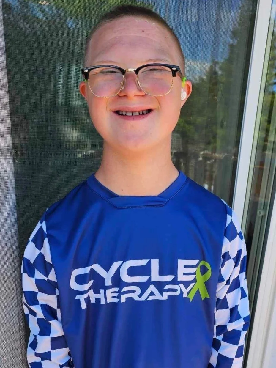 Young person smiling wearing glasses and a blue cycling therapy jersey with checkered sleeves, standing in front of a screen door or window.