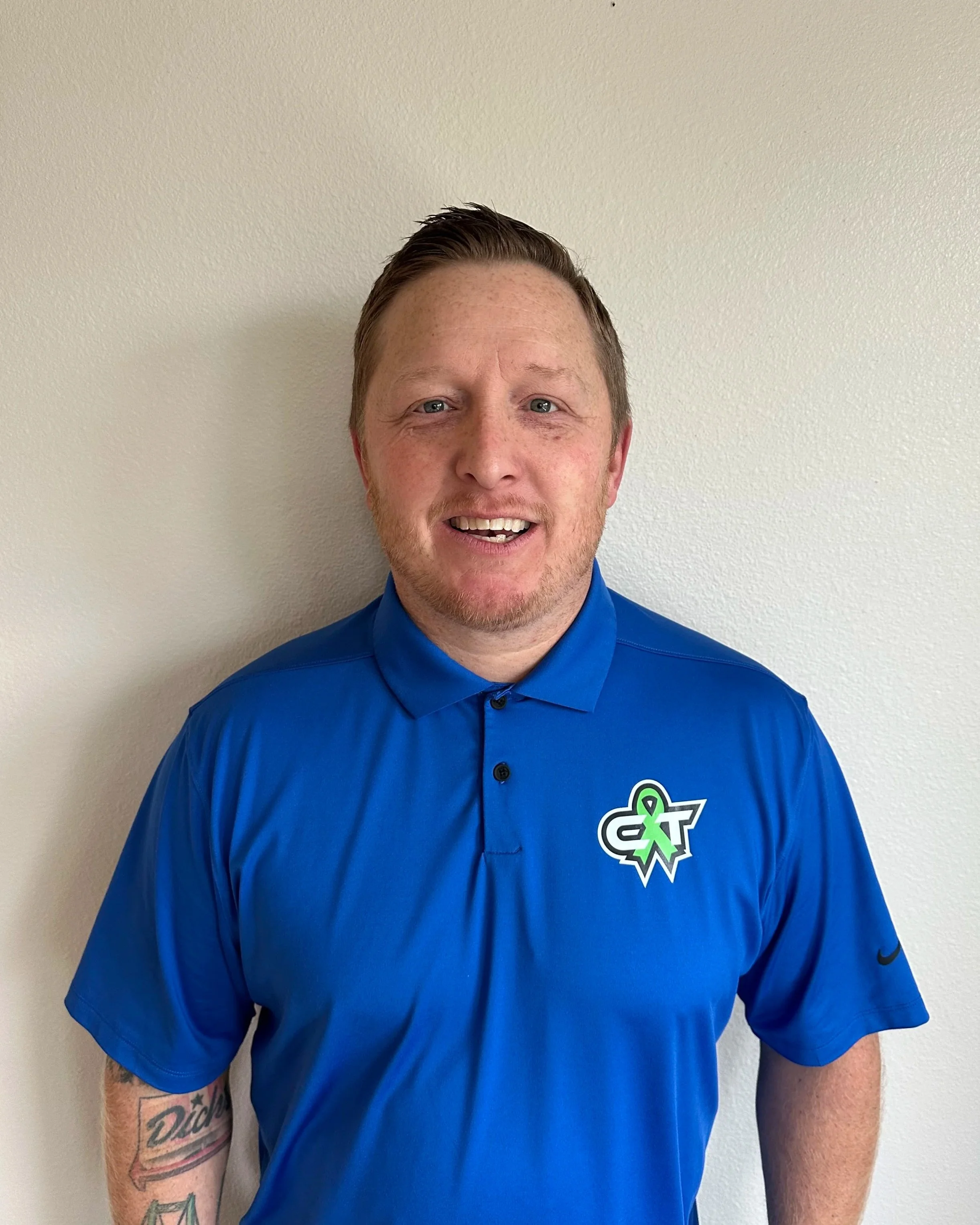 A man with light skin, short light brown hair, and a light beard, smiling and wearing a blue polo shirt with a logo on it, standing against a plain beige wall.