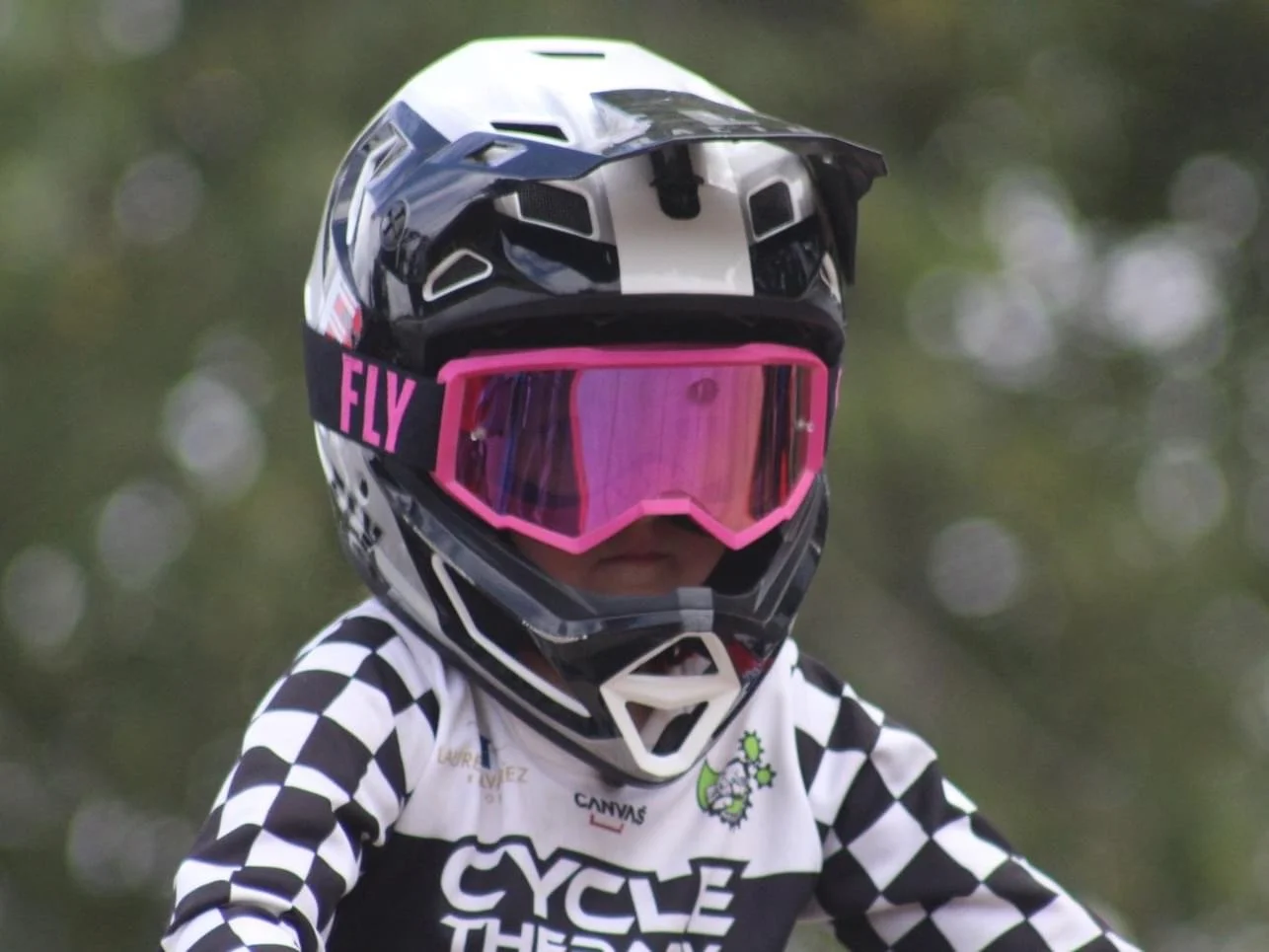 Close-up of a BMX racer, biker wearing a black and white checkered jersey, pink goggles, a white and black helmet with vents, and a helmet strap with the word 'FLY' in pink, outdoors with blurred green trees in the background.