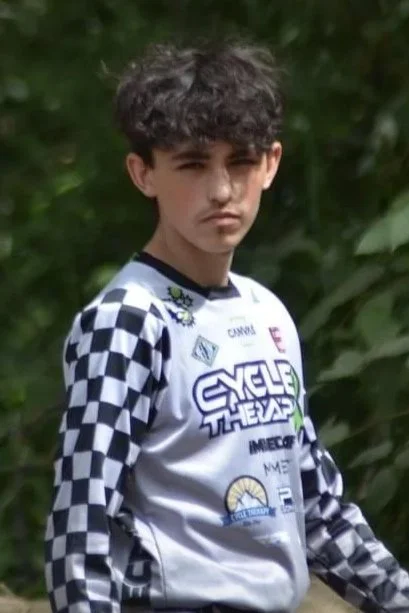 Young boy with curly hair standing outdoors in front of green foliage, wearing a racing-style jersey with checkered sleeves and various logos.