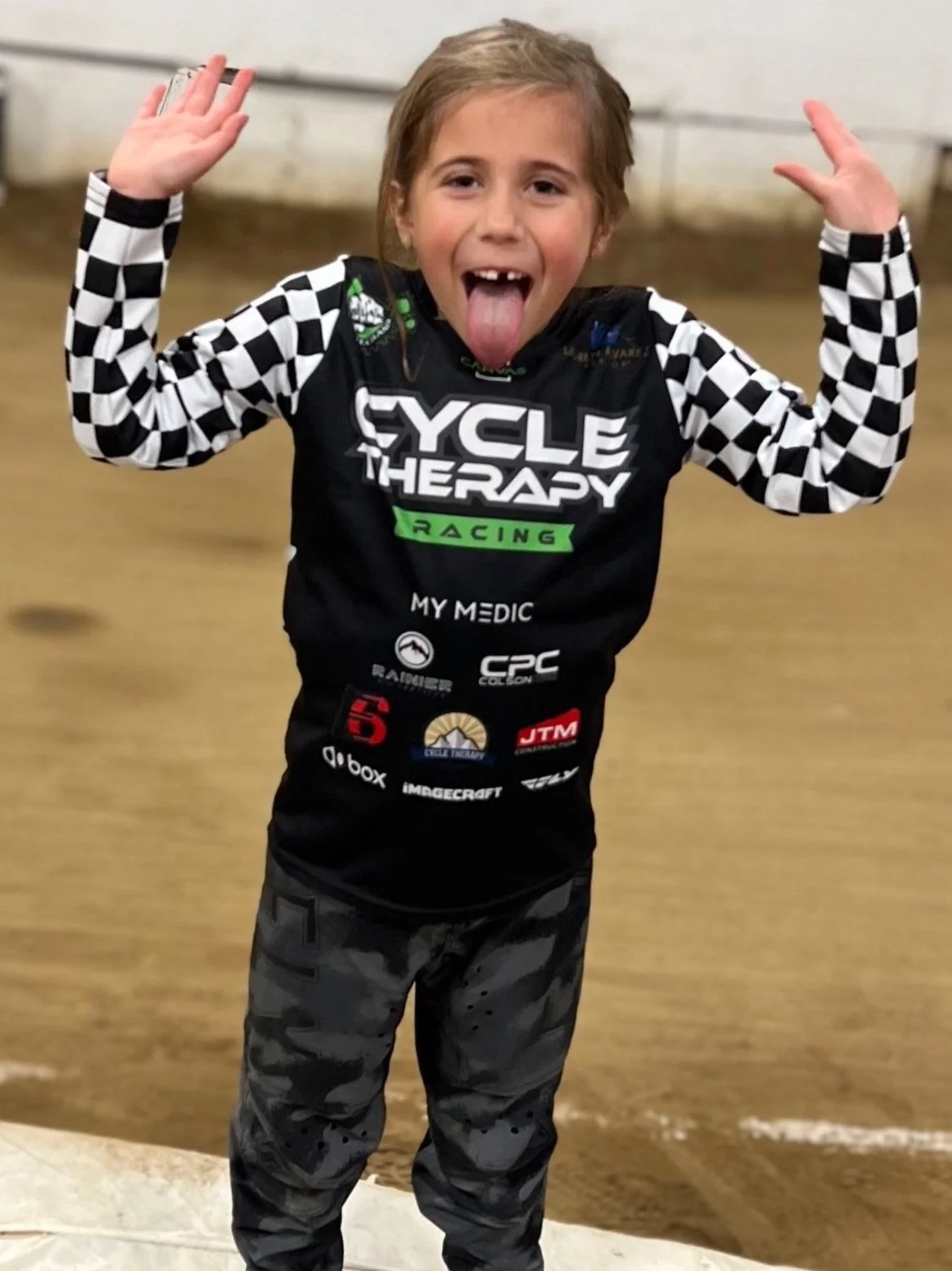 A young girl posing with her tongue out and hands raised, wearing a black and white checkered long-sleeve shirt under a black cycling jersey with various sponsor logos, standing on an indoor dirt track.