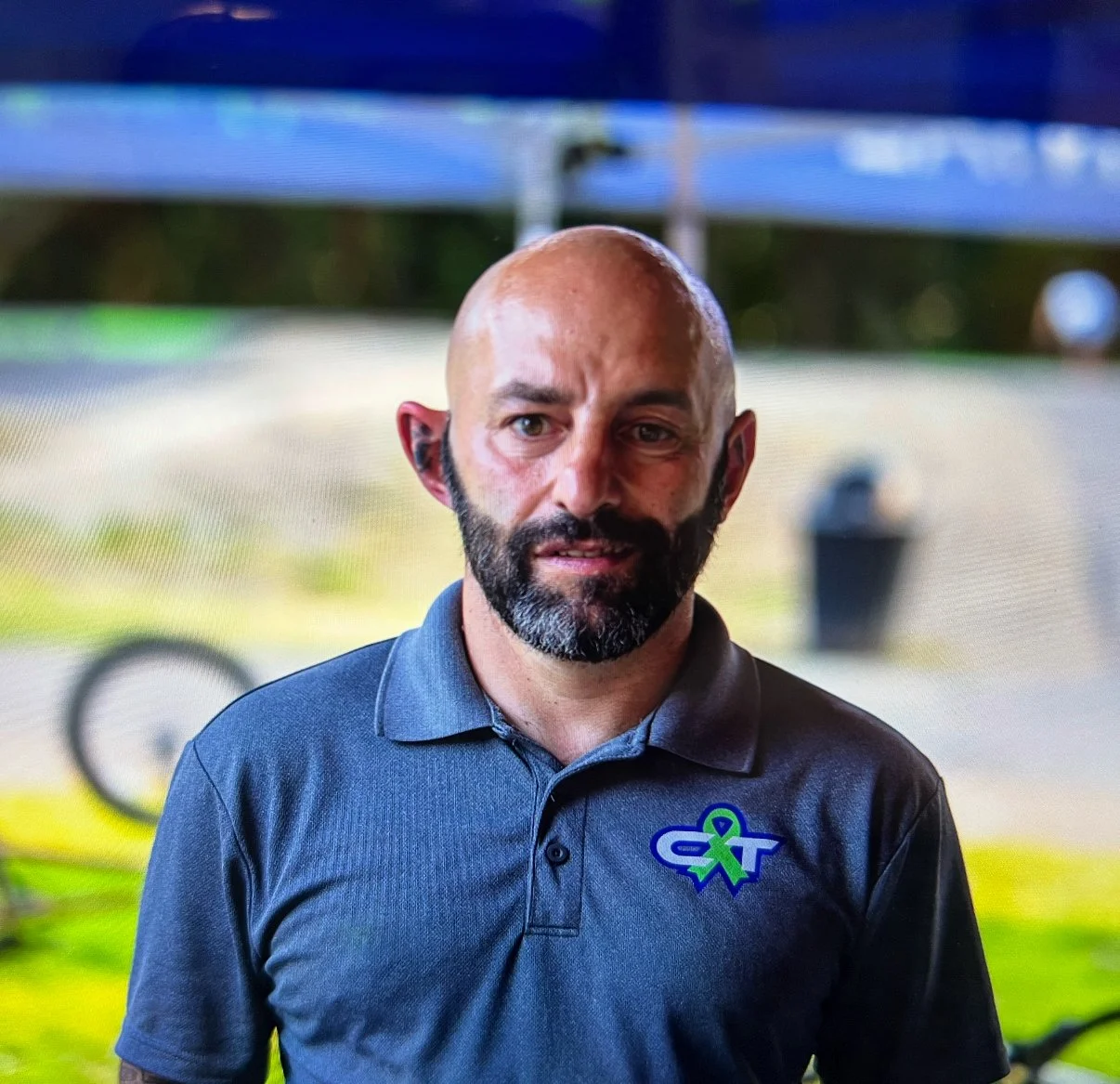 A bald man with a beard wearing a dark gray polo shirt with a logo on the chest, standing outdoors with a blurred background of trees, a bicycle, and a trash can.