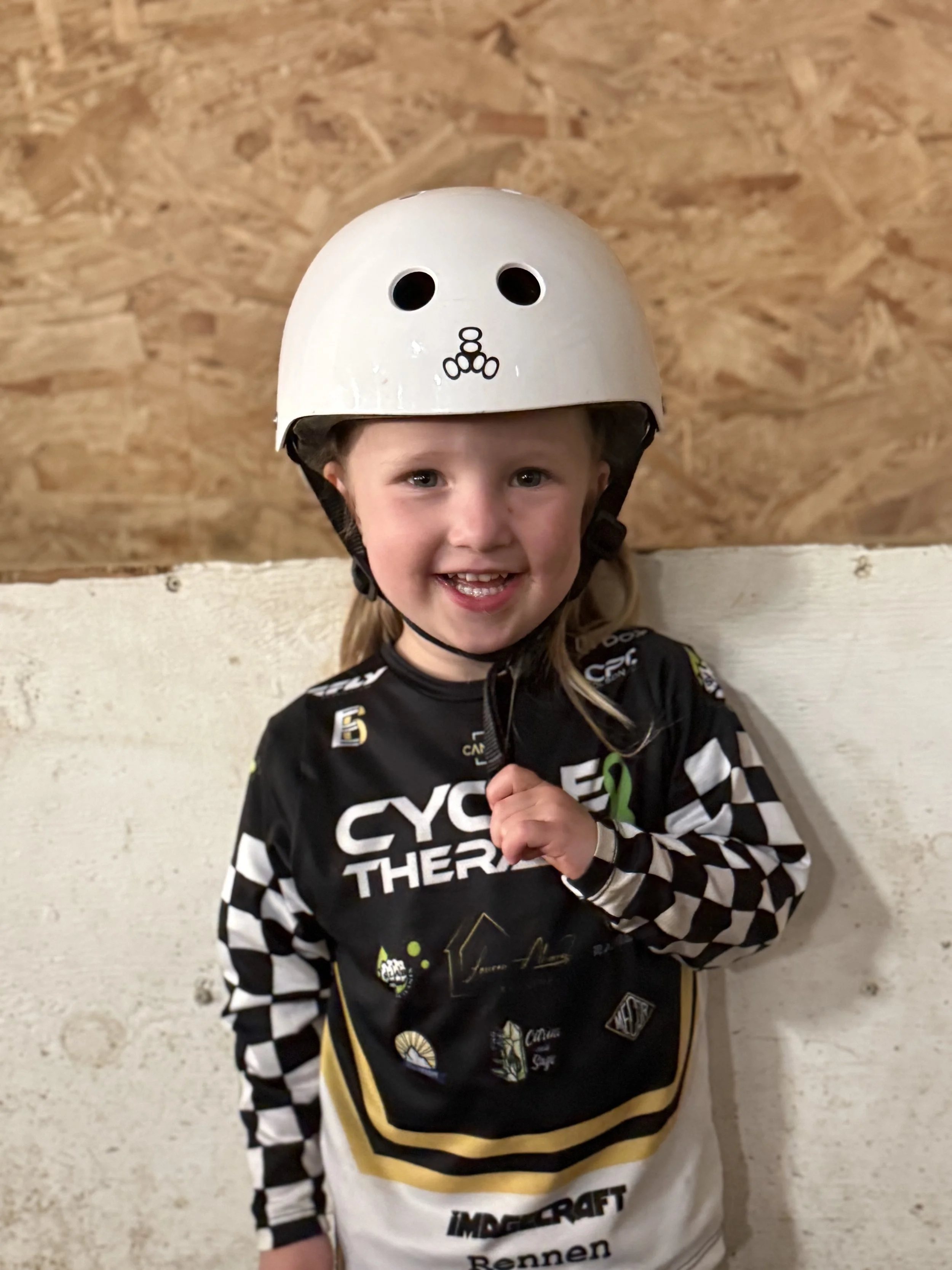 A young girl smiling while wearing a black and white checkered racing jersey and a white helmet with a face design.