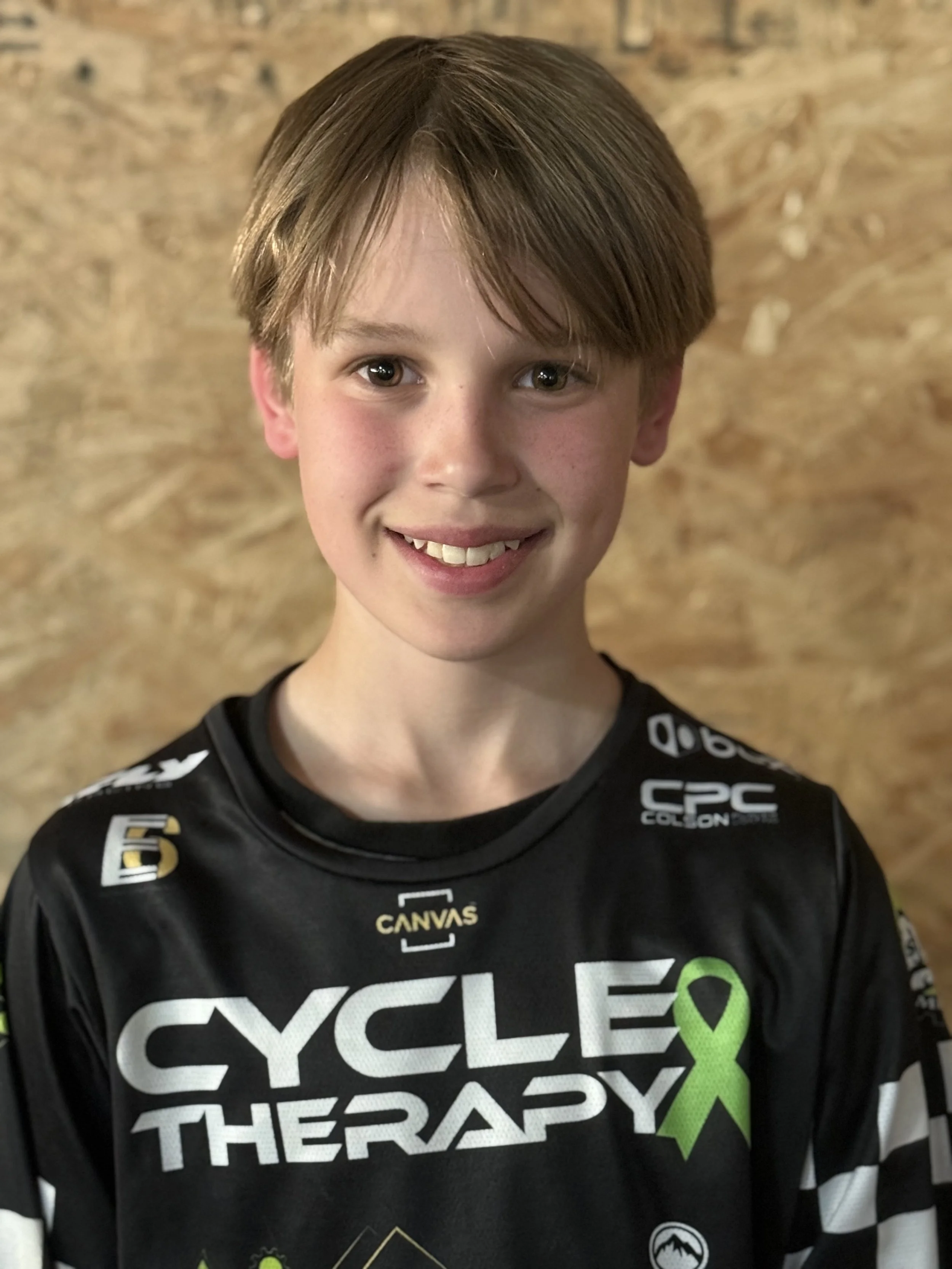 A young boy with light brown hair and a bright smile, wearing a black cycling jersey with logos and a lime green awareness ribbon, standing in front of a wooden wall.