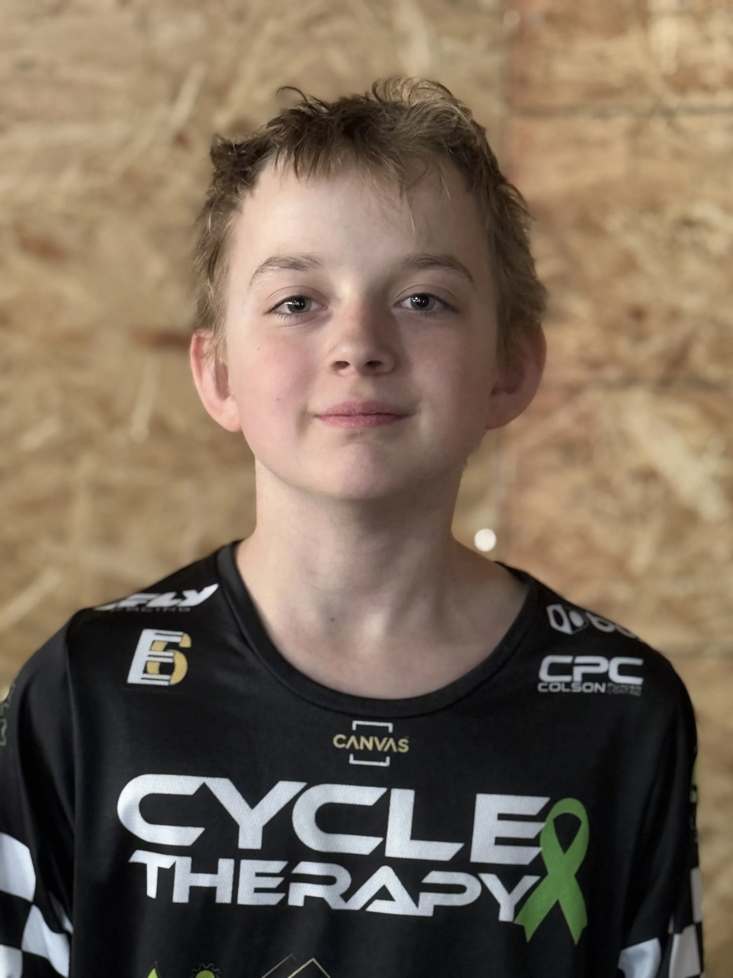 A young boy with light brown hair and blue eyes, wearing a black cycling jersey with logos and the words "Cycle Therapy" and a green ribbon, standing against a wooden background.