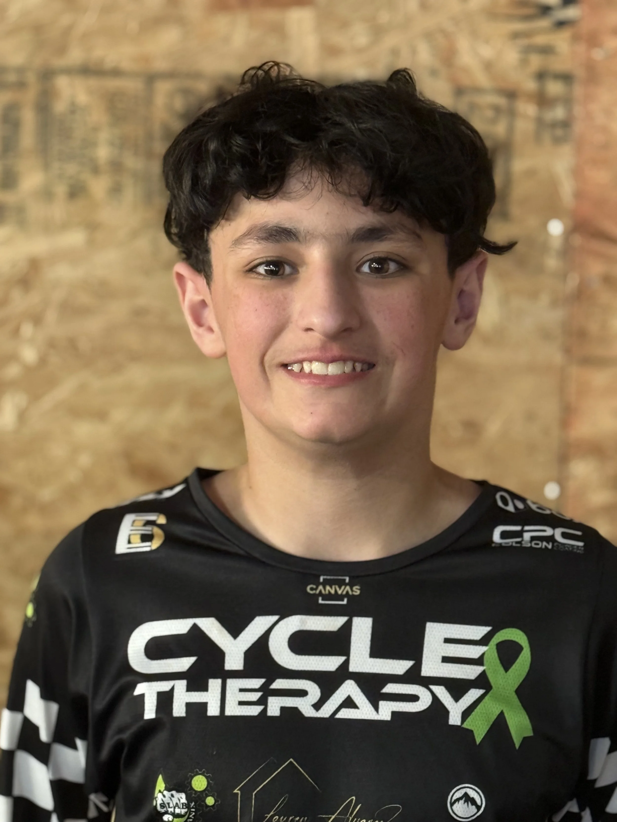 A young person with curly dark hair smiling at the camera, wearing a black cycling T-shirt with logos and a green ribbon, standing in front of a wooden background.
