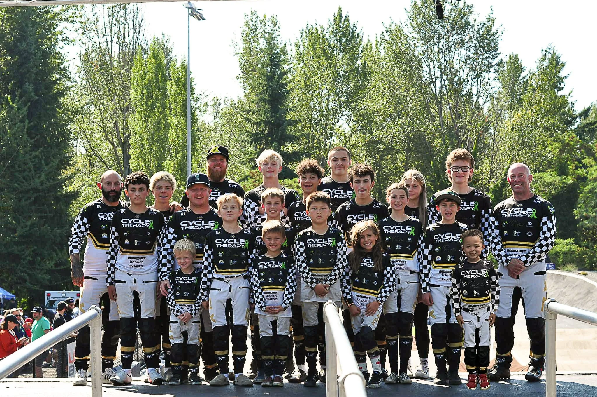 Group of young motocross racers in black and white checkered racing suits and helmets posing outdoors on a sunny day with trees in the background.