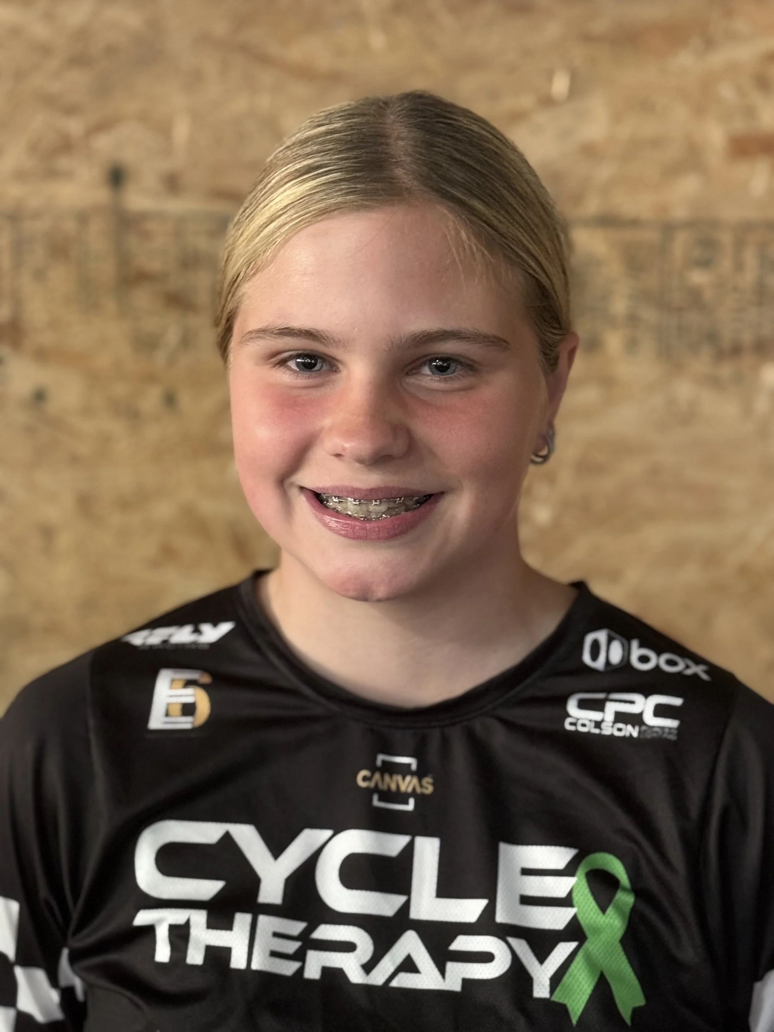 A smiling young girl with blonde hair in a ponytail wearing a black cycling jersey with white and green lettering and logos, standing in front of a wooden background.