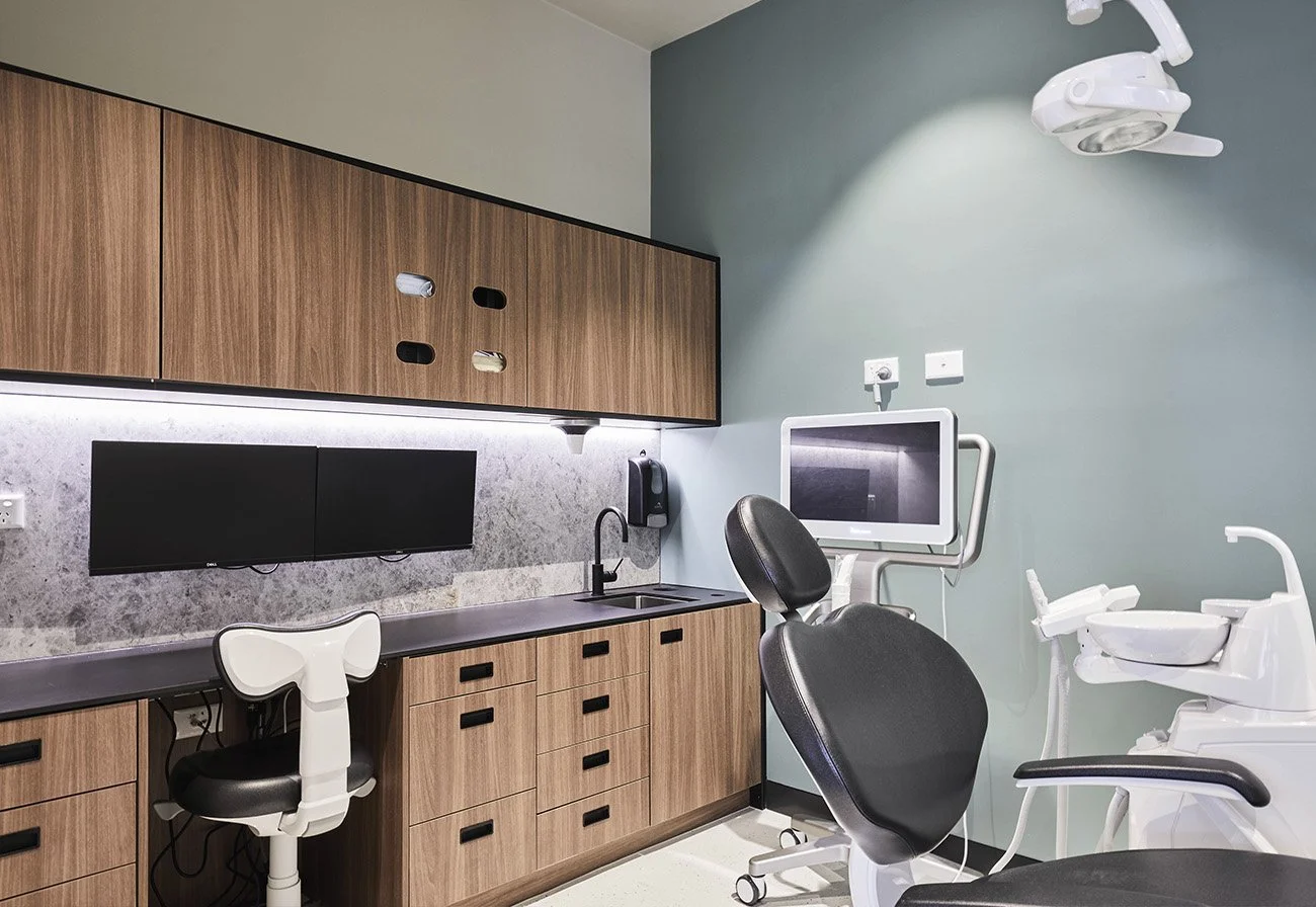 A modern dental examination room with dental chair, monitor, overhead light, cabinetry, and sink.