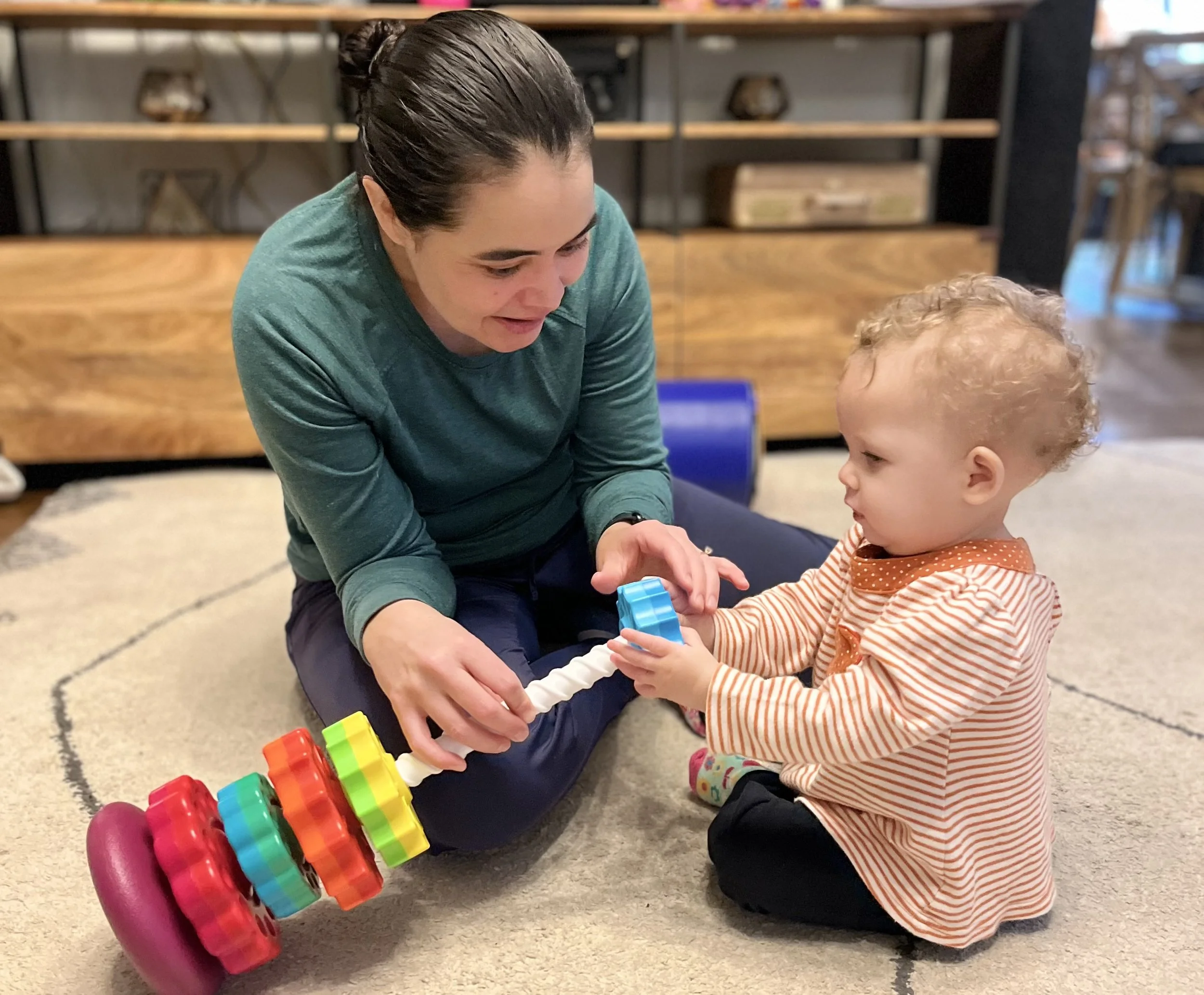A woman in a teal sweatshirt and a young child with curly blonde hair, wearing a striped orange and white shirt, sitting on a beige carpeted floor, playing with colorful stacking rings. The woman is smiling and engaging with the child in a room with wooden furniture and various items in the background.