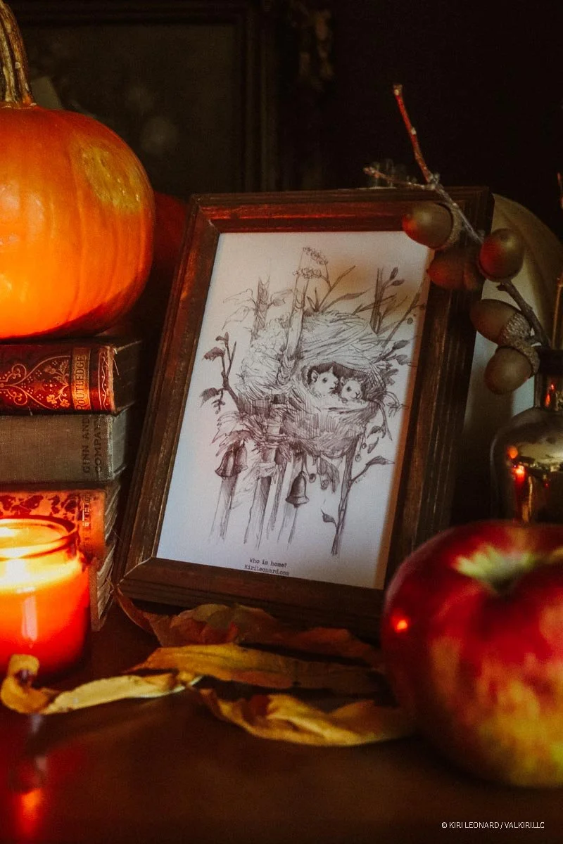 A framed sketch of two owls in a nest, surrounded by tree branches, with a small bell hanging below the nest. The scene is set among candles, an apple, a pumpkin, and books on a dark surface.