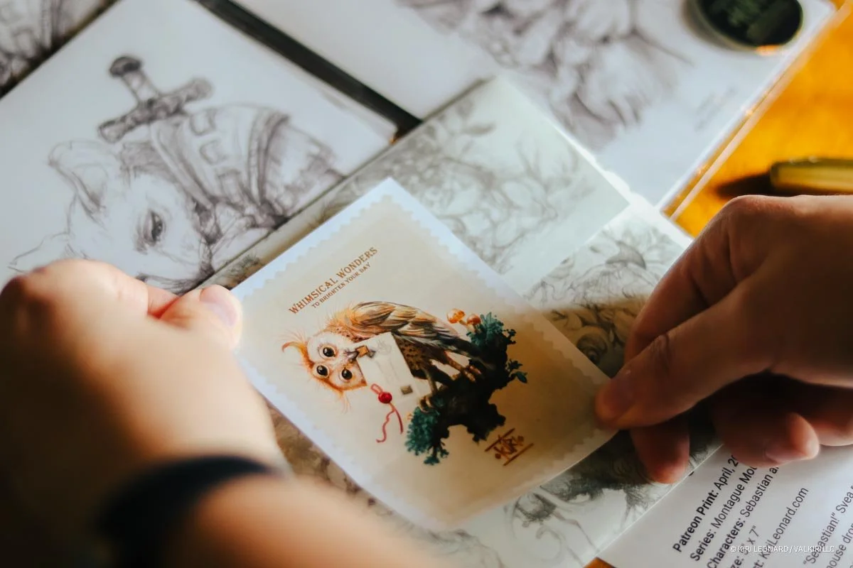 Person peeling a sticker featuring a whimsical owl with a bow and flowers from a sheet of paper in an art or sketchbook.
