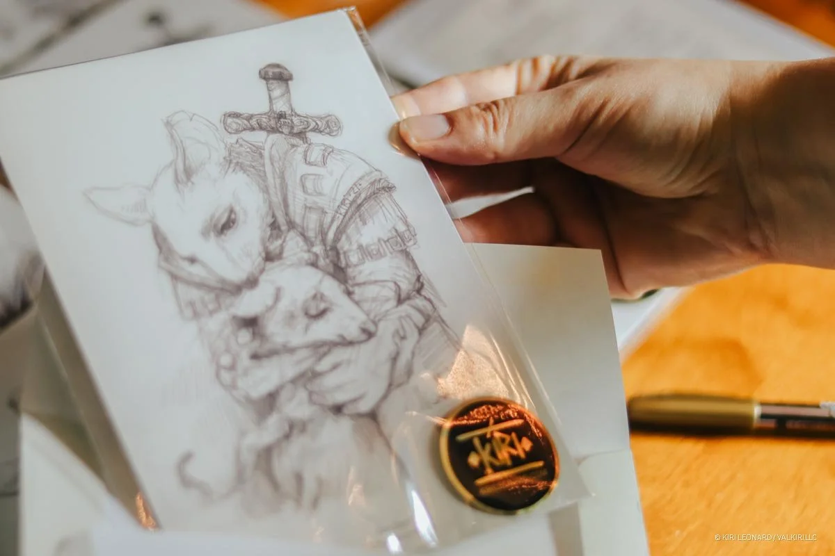 A hand holding a small sketch of two dogs, one of which has a sword behind its head, in front of a table with a black pen and a round badge or button.