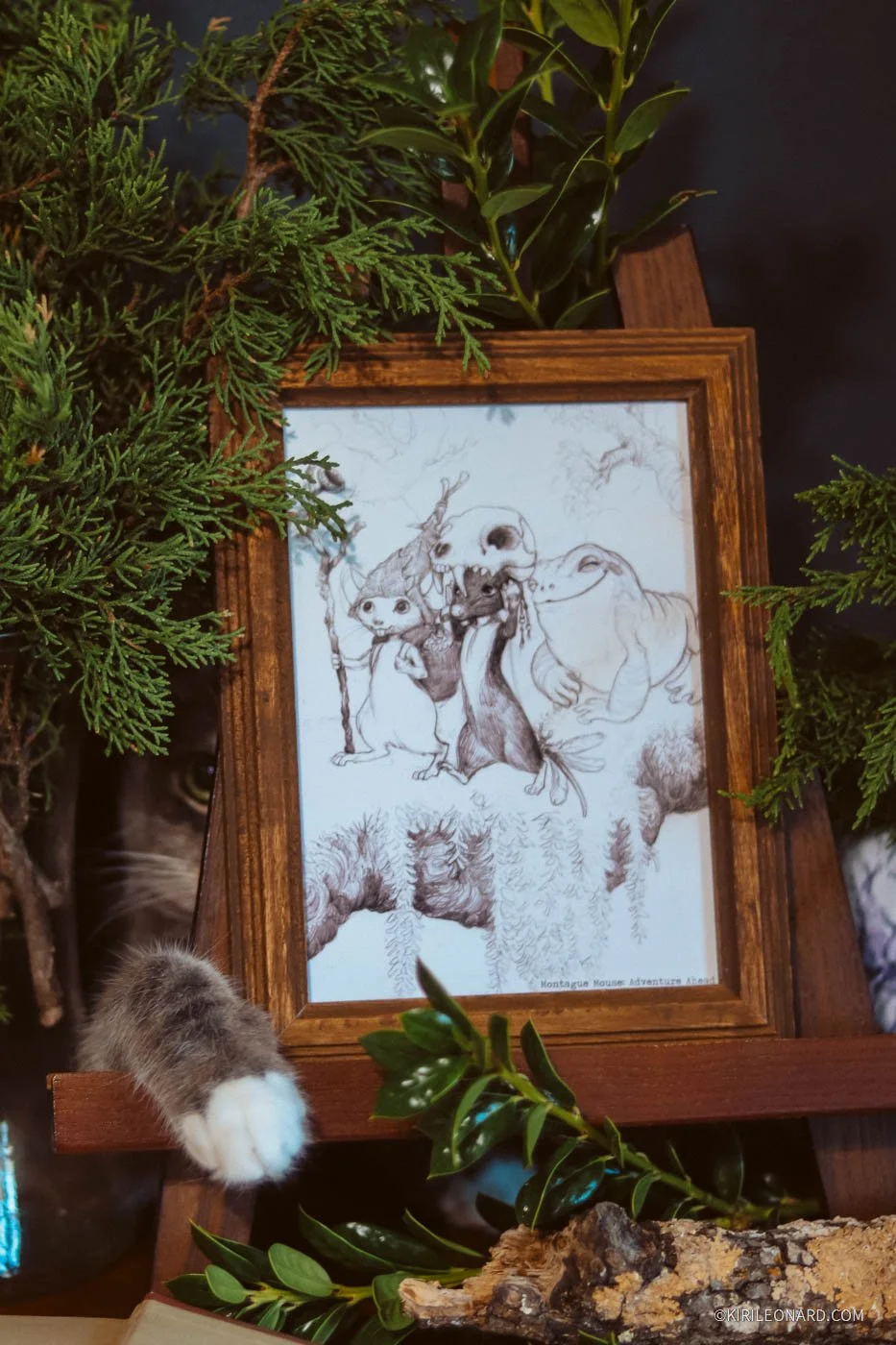 A framed sketch of animated characters including a girl, a large creature with a skull face, a dinosaur, and a wizard, surrounded by green plants and branches, with a cat's paw in the foreground.