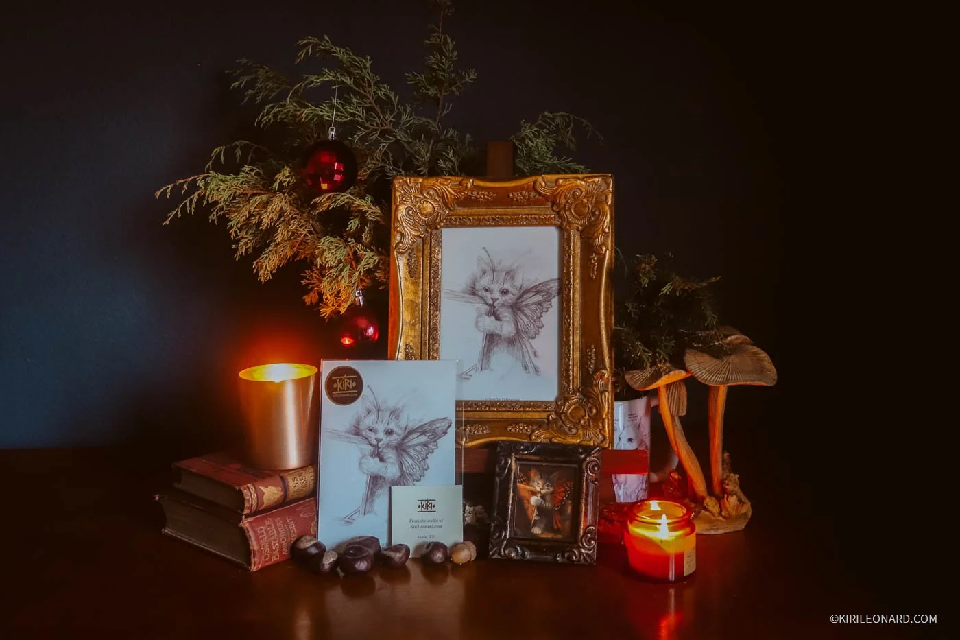 A framed sketch of a fairy cat, Christmas decorations including red ornaments, a candle, books, and a small framed picture of the same fairy cat sketch with a candle in front, all arranged on a dark surface.