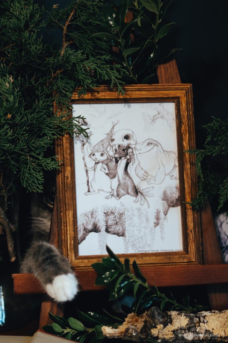 A framed drawing of animated mole characters is decorated with green foliage and a cat paw is visible in the lower left corner.