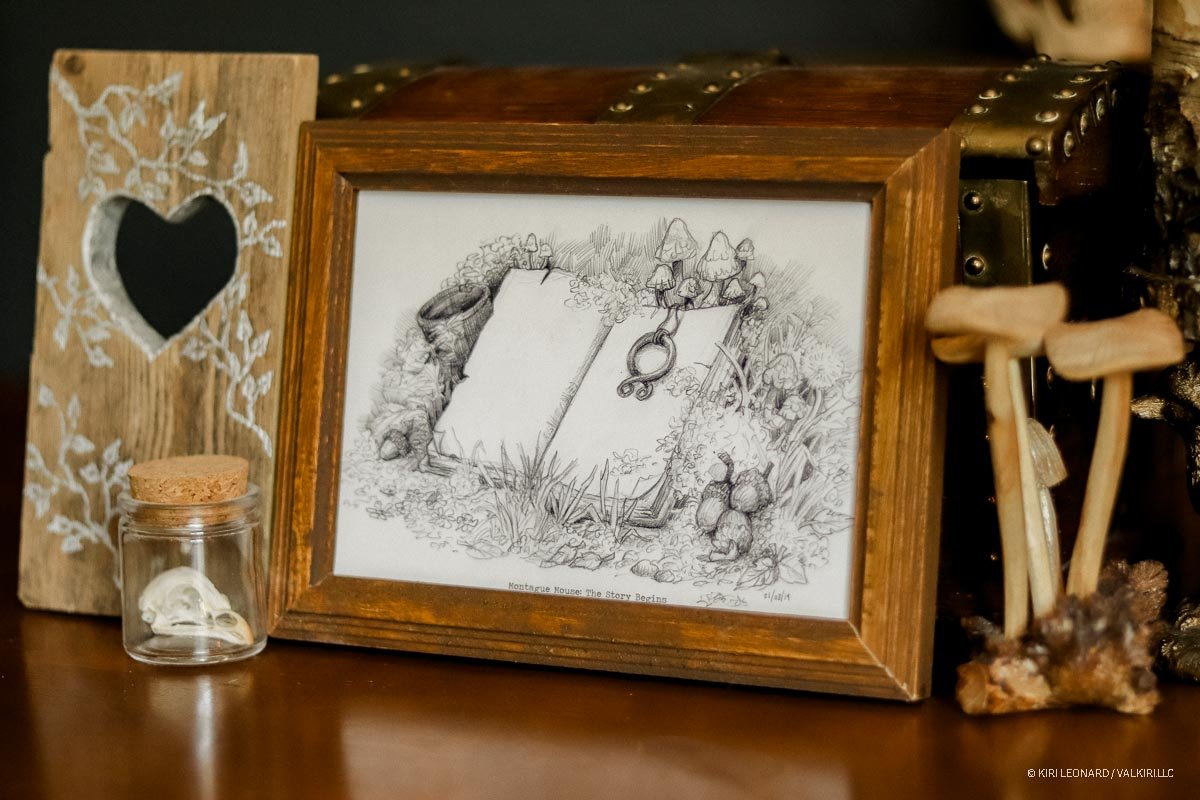 A framed black-and-white drawing titled 'Montague Mouse: The Story Begins' features a scene with an open book surrounded by mushrooms, plants, and small creatures resembling mice. To the left, there is a wooden decorative piece with a heart-shaped ho