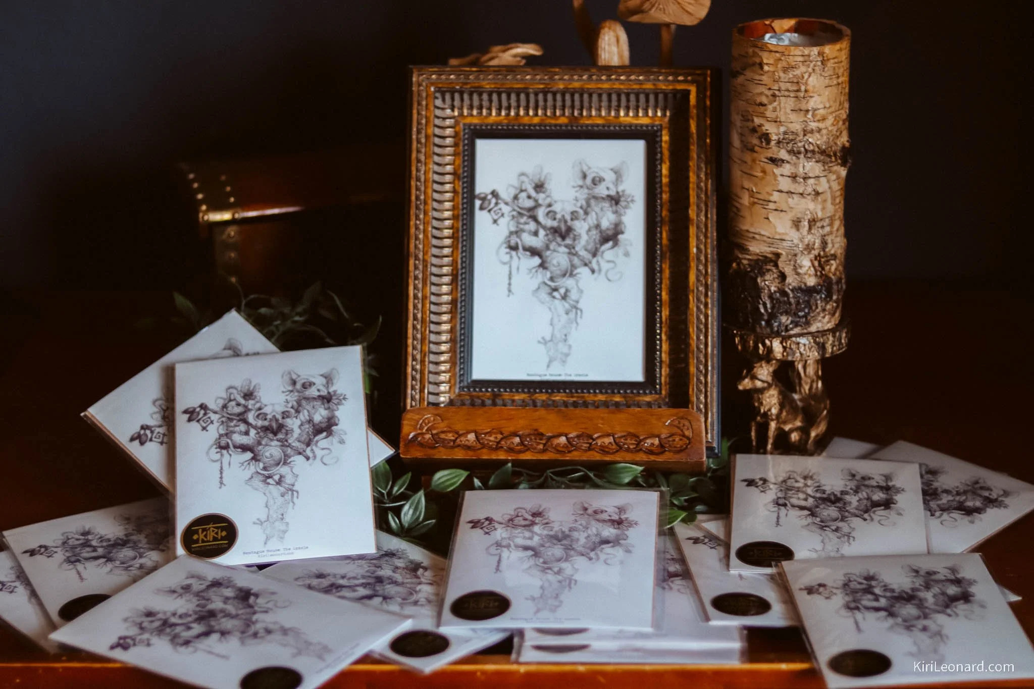 Display of prints and cards featuring a line art drawing of a cherubic figure with wings, surrounded by decorative elements. The display includes a framed print, multiple smaller prints or cards laid out on a surface, a decorative wooden stand, and a
