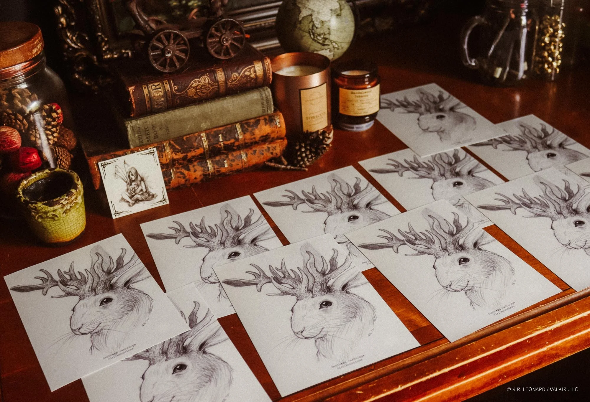Drawings of a deer with large antlers, arranged on a wooden table among books, candles, a globe, and decorative items in a cozy setting.